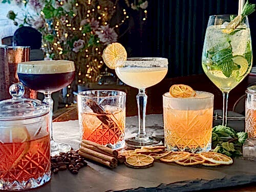 Hurley House - A selection of colorful cocktails garnished with citrus slices, mint, and cinnamon sticks, displayed on a slate tray with dried fruit and fairy lights in the background.