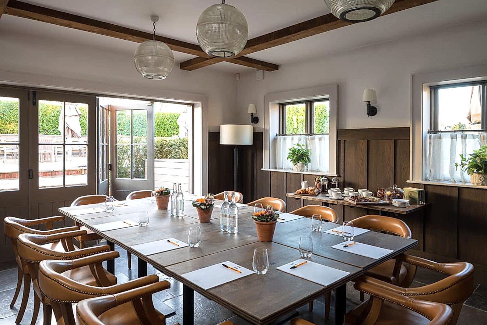 Hurley House - A cozy meeting room with a large wooden table surrounded by leather chairs. The table is set with placemats, glasses, and water bottles. Natural light streams through open double doors, and potted plants decorate the space.