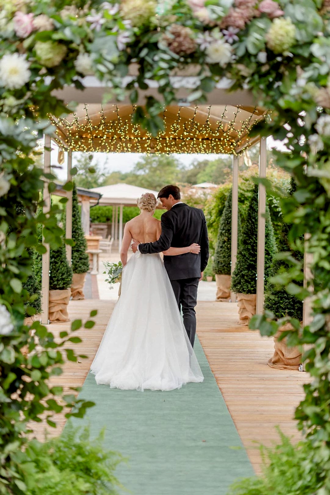 Hurley House - A bride in a white gown and a groom in a dark suit walk arm-in-arm down a green carpeted aisle at a boutique wedding venue in Berkshire, surrounded by lush greenery and soft string lights, creating a romantic garden wedding setting.