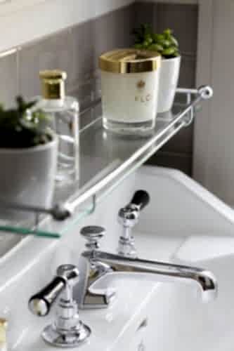Hurley House - A bathroom sink with chrome faucets. Above it, a glass shelf holds a candle labeled FLO, a small potted plant, and a perfume bottle. The background features beige tiles.