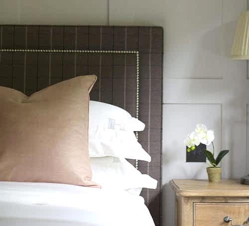 Hurley House - A cozy bedroom corner featuring a bed with white linens and a large brown pillow. Theres a dark checkered headboard and a wooden nightstand holding a small white orchid in a pot and a lamp. The walls are paneled, adding elegance to the space.