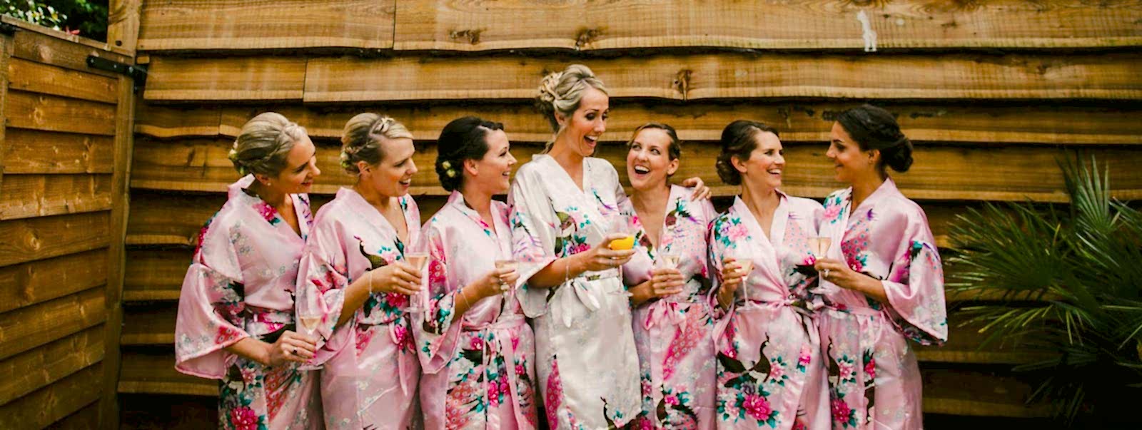 Hurley House - A group of seven bridesmaids in floral robes, six in pink and one in white, stand together laughing and holding drinks by a wooden fence. They appear to be celebrating at a luxury wedding venue in Berkshire, possibly during a pre-wedding event.