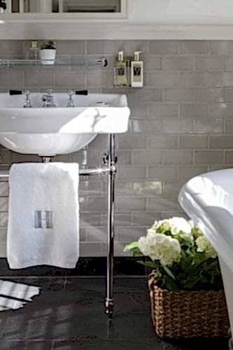 Hurley House - A bathroom with a white sink on chrome legs, set against a beige subway tile wall. A small plant is on a glass shelf above the sink, and a wicker basket with white hydrangeas sits on the floor. A white towel with an H monogram hangs from the sink.