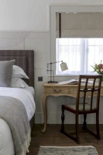 Hurley House - A cozy bedroom corner with a neatly made bed featuring a gray headboard and white linens. Adjacent is a wooden desk with an angled lamp and an open book, accompanied by a wooden chair. A window with sheer curtains lets in soft light.