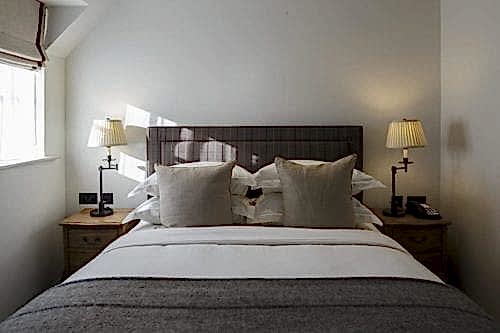 Hurley House - A cozy bedroom with a neatly made double bed, featuring a checkered headboard and two beige pillows. Two bedside tables hold matching lamps. Soft light comes through a window on the left, creating a warm atmosphere.