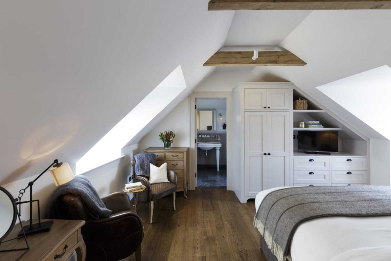 Hurley House - Cozy attic bedroom with a white wardrobe, wooden dresser, armchair, and a desk lamp. A bed with a gray blanket is on the right. Sloped ceiling with exposed wooden beams and a skylight. A bathroom is visible through an open door in the background.