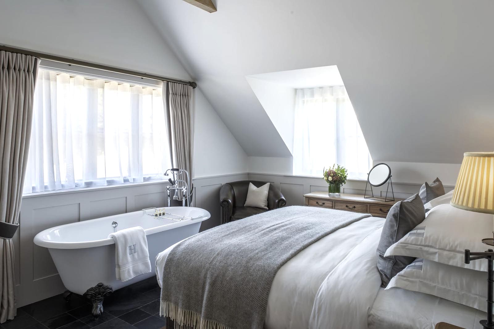 Hurley House - A cozy bedroom with a clawfoot bathtub by the window, a large bed with gray bedding, a wooden dresser with a mirror and vase of flowers, soft lighting from a table lamp, and slanted ceilings with a skylight.