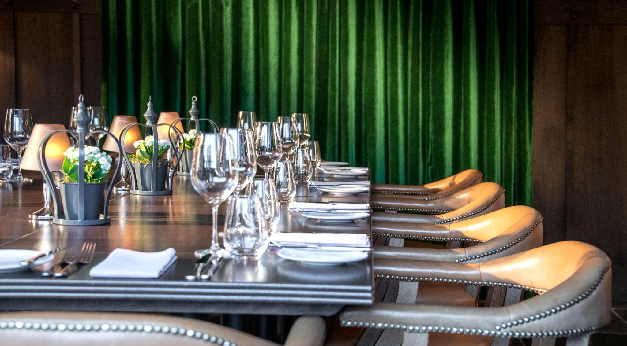 Hurley House - A elegantly set dining table with brown leather chairs, wine glasses, and potted plants, against a backdrop of lush green curtains.