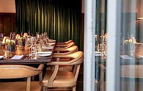 Hurley House - A well-set dining table in an elegant restaurant with wooden chairs and a green velvet curtain in the background. The table is adorned with wine glasses, white napkins, and small decorative flowers, creating a sophisticated atmosphere.