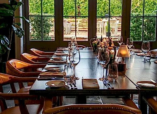 Hurley House - A long wooden dining table is elegantly set with plates, silverware, wine glasses, and small lamps. Leather chairs are neatly arranged around the table. Large windows in the background provide a view of greenery outside.