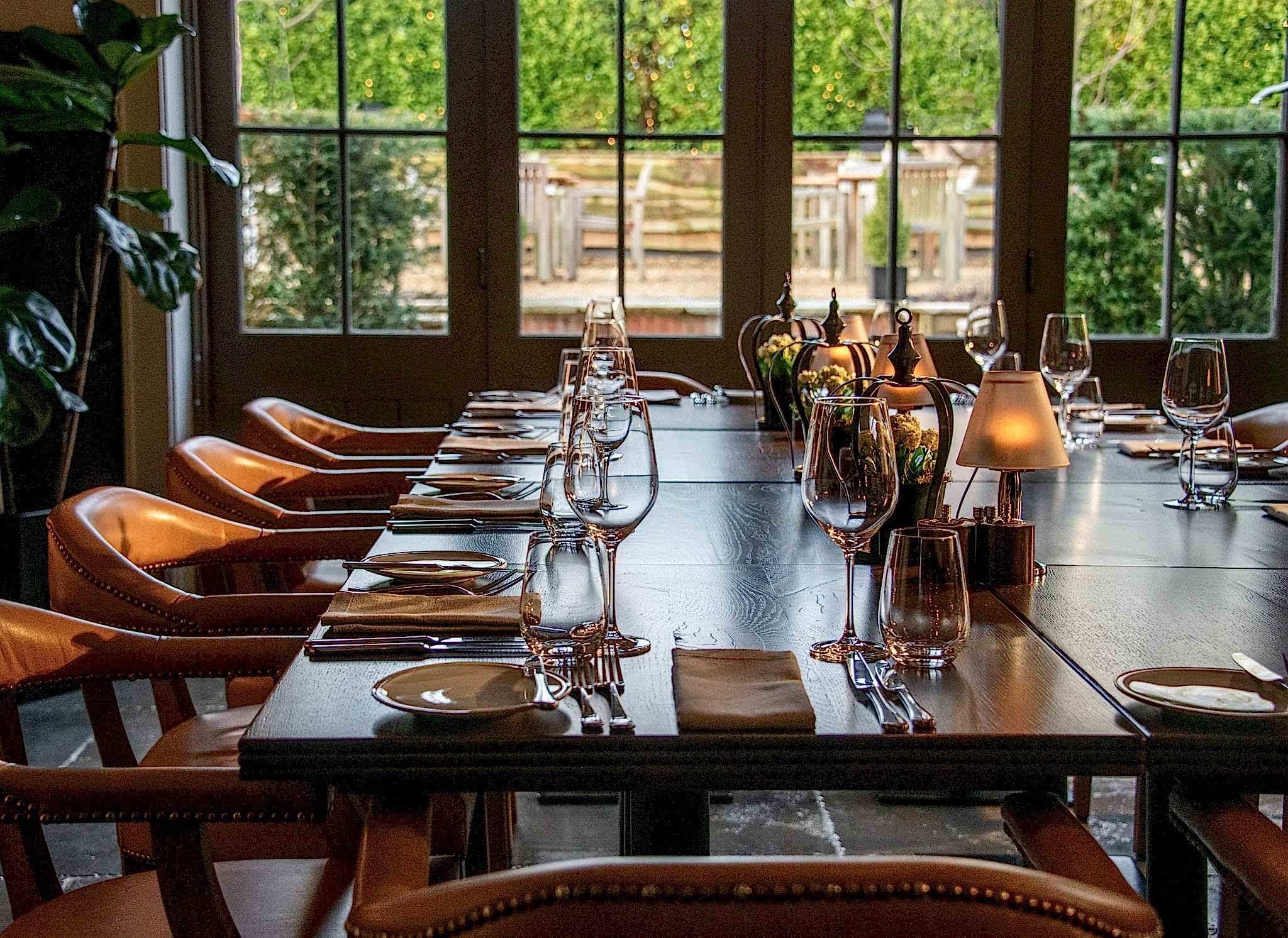 Hurley House - A long wooden dining table is elegantly set with plates, silverware, wine glasses, and small lamps. Leather chairs are neatly arranged around the table. Large windows in the background provide a view of greenery outside.