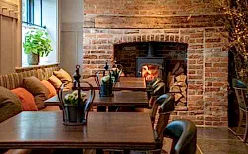 Hurley House - Cozy cafe with a rustic brick fireplace, a wood-burning stove, and stacked logs. Wooden tables with small potted plants and cushioned seating line the wall. Warm lighting adds to the inviting atmosphere.