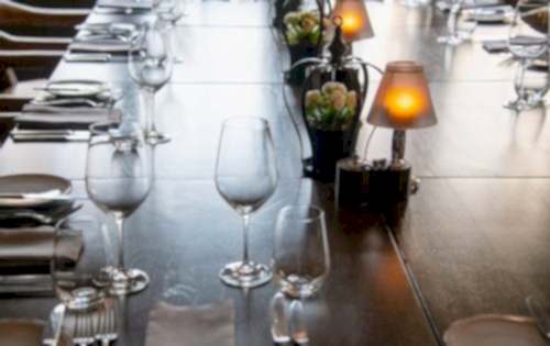 Hurley House - A long wooden table set for dining, with neatly arranged wine glasses, plates, and cutlery. Decorative lamps and small vases with flowers line the center, creating an elegant and cozy atmosphere.