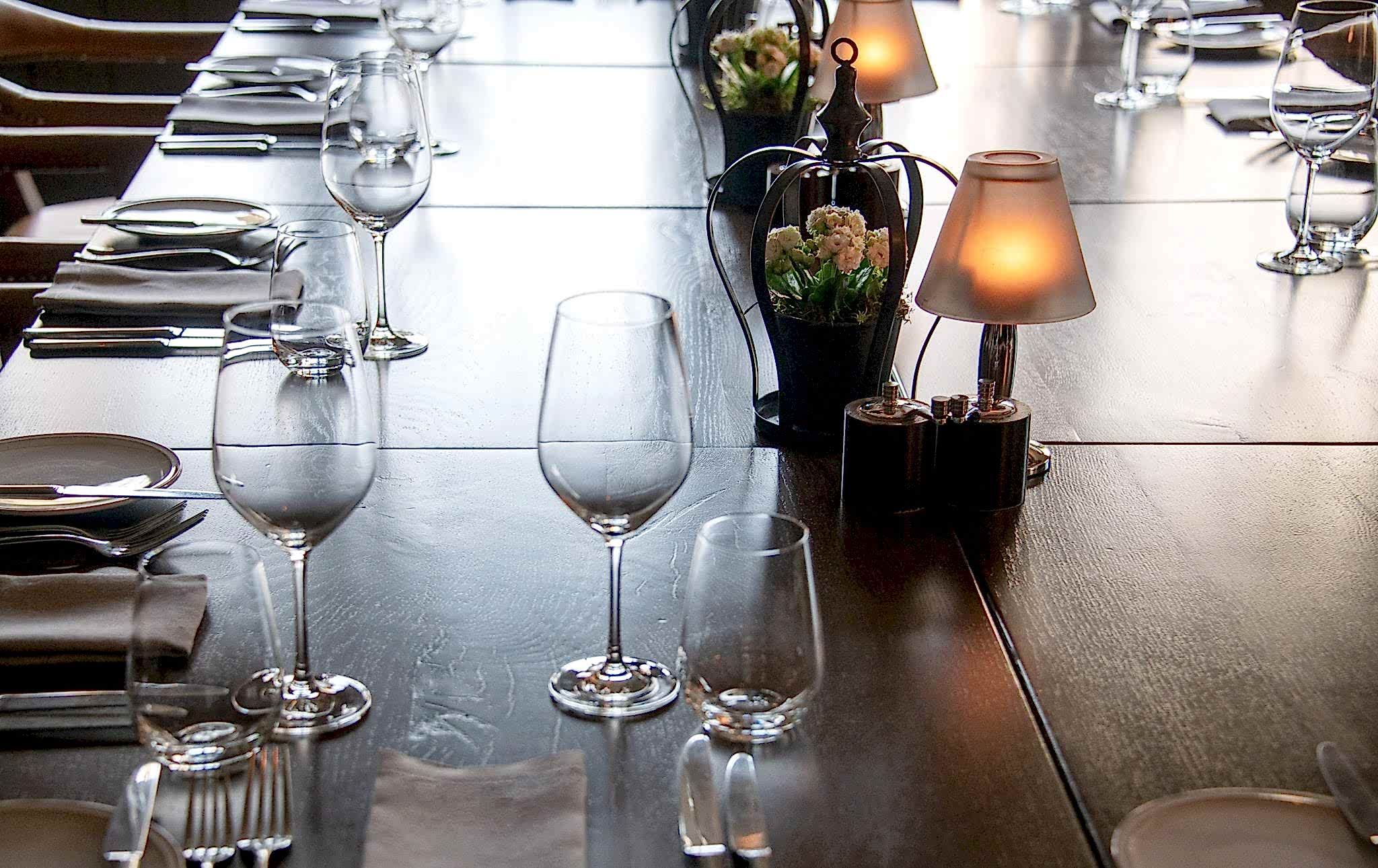 Hurley House - A long wooden table set for dining, with neatly arranged wine glasses, plates, and cutlery. Decorative lamps and small vases with flowers line the center, creating an elegant and cozy atmosphere.