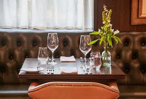 Hurley House - A neatly set dining table for two with wine glasses, water glasses, and cutlery on a dark wooden table. A small vase with white flowers decorates the table. The background features tufted leather seating and a curtain.