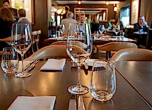Hurley House - A dimly lit restaurant with elegantly set tables, featuring empty wine glasses and folded napkins. Patrons are seen in the background, creating a cozy and inviting ambiance.