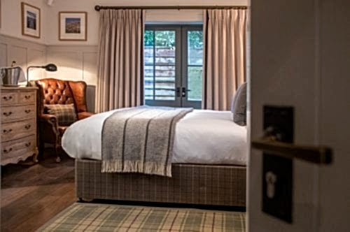 Hurley House - Cozy bedroom with a large bed, gray blanket, and plaid rug. A brown leather armchair and a wooden dresser are on the left. Soft light filters through curtains on a glass door, revealing a wooden fence outside. A lamp sits on the dresser.