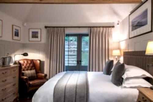 Hurley House - A cozy bedroom with a large bed adorned with gray pillows and a blanket. A brown leather armchair is placed beside a wooden dresser. Framed pictures hang on white-paneled walls. Soft lighting filters through closed beige curtains on a glass door.