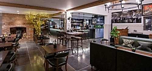 Hurley House - A modern, cozy restaurant interior with brick walls, a fireplace, tables, chairs, and a central bar. There are plants, decorative lighting, a wooden beam ceiling, and framed pictures on the wall, creating a welcoming atmosphere.