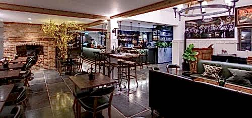 Hurley House - A cozy, modern bar and restaurant interior with wooden beams, a brick fireplace, and various seating arrangements. Green plants and framed photos add decoration. The bar area is well-lit and stocked with bottles.