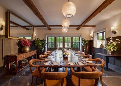 Hurley House - A warmly lit dining room features a long table set for a meal with elegant settings. Leather chairs surround the table, and large windows show an outdoor view. Wooden beams and plants add to the cozy ambiance.