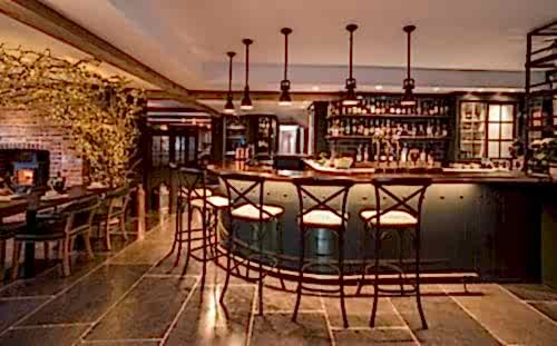 Hurley House - A cozy, warmly lit bar with high stools, a wooden counter, and shelves stocked with bottles. Rustic decor includes a brick fireplace, a stone floor, and ambient lighting, creating an inviting atmosphere. A dining table with chairs is partially visible.