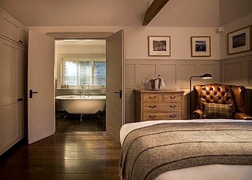 Hurley House - A cozy bedroom with a plush bed, a brown leather chair, and a wooden dresser. The room is warmly lit, revealing a white bathtub in a connected bathroom. Framed pictures hang on the walls, and a tray with a teapot and cups sits on the dresser.