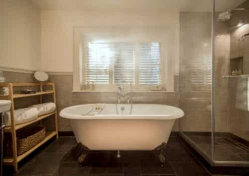 Hurley House - A cozy bathroom with a freestanding white bathtub on clawfoot legs, a wooden shelf with towels, a glass-enclosed shower, and a window with shutters. The room has beige tile walls and a dark tiled floor.
