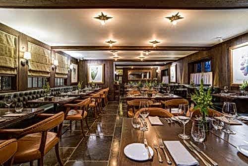 Hurley House - A cozy and elegantly furnished restaurant interior featuring wooden tables, leather chairs, and soft lighting. The tables are set with plates, cutlery, and glasses, surrounded by artwork and large windows with blinds.