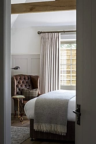 Hurley House - A cozy bedroom with a large window covered by beige curtains. A brown leather armchair sits in the corner next to a small round table with a lamp. A neatly made bed with a gray blanket is visible, and a wooden beam runs across the ceiling.