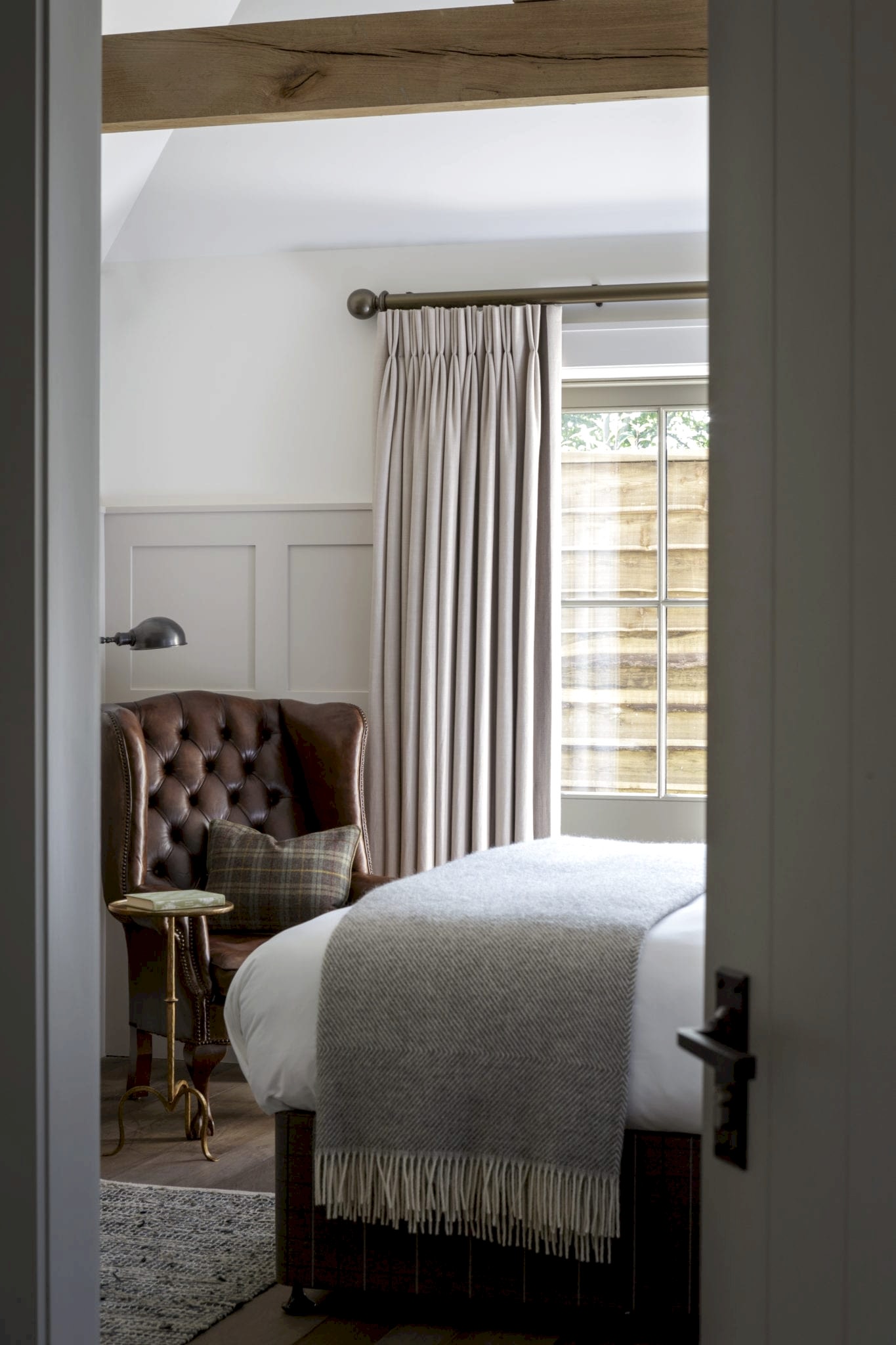 Hurley House - A cozy bedroom with a large window covered by beige curtains. A brown leather armchair sits in the corner next to a small round table with a lamp. A neatly made bed with a gray blanket is visible, and a wooden beam runs across the ceiling.