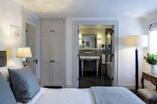 Hurley House - A cozy bedroom with a neatly made bed featuring gray pillows and a blanket. Theres a bedside lamp, a wardrobe, and an open door leading to a bathroom with a sink and mirror. Natural light filters through a window with a plant on a nearby table.