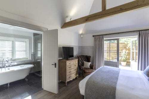 Hurley House - A cozy bedroom featuring a bed with gray and white linens, a wooden dresser with a TV, and a comfortable armchair. The room has an open bathroom with a freestanding bathtub and a door leading to a patio with wooden fencing and greenery.