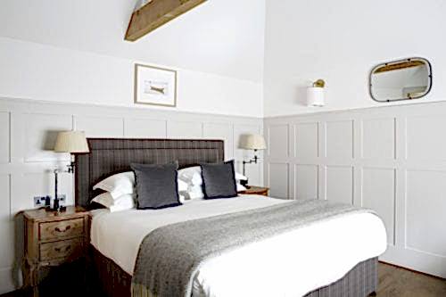 Hurley House - A cozy bedroom with a double bed featuring a checkered headboard, white and gray bedding, and two gray pillows. Two wooden nightstands with lamps flank the bed. A framed picture, an oval mirror, and a wall sconce decorate the white paneled walls.
