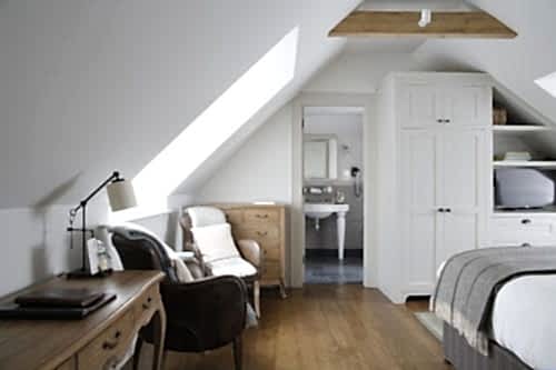 Hurley House - Cozy attic bedroom with slanted ceilings, featuring a wooden floor, white wardrobe, armchair, desk with lamp, and a view into a small bathroom. Natural light streams in through skylights, creating a serene atmosphere.