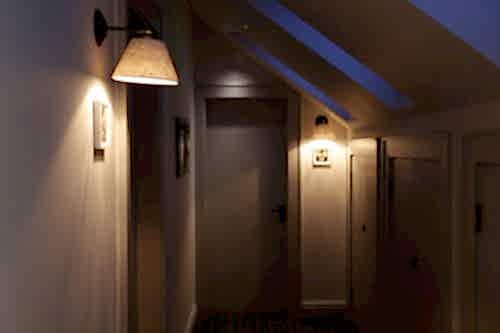 Hurley House - A dimly lit hallway with a sloped ceiling features wall lamps casting warm light. The walls are adorned with small framed pictures, and there are several wooden doors along the hallway.
