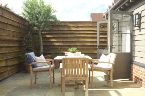 Hurley House - Outdoor patio with wooden furniture, including a table and two chairs, on stone tiles. Potted plants line the wooden fence. An open door leads inside. The scene is bright and inviting under a clear sky.