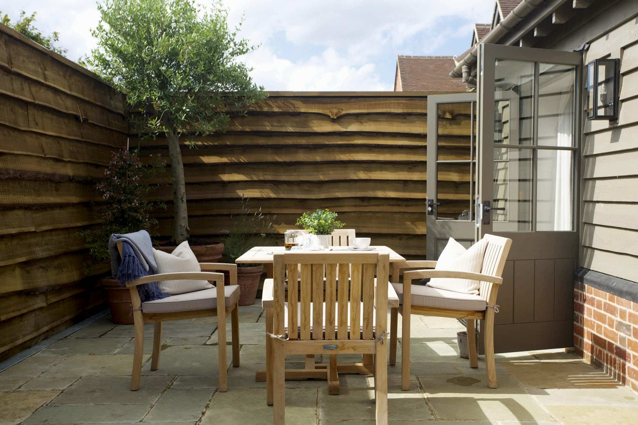 Hurley House - Outdoor patio with wooden furniture, including a table and two chairs, on stone tiles. Potted plants line the wooden fence. An open door leads inside. The scene is bright and inviting under a clear sky.