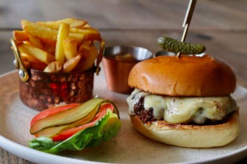 Hurley House - A cheeseburger with lettuce, tomato, and pickles is served on a plate. A small pickle is skewered on top. A side of crispy fries is in a copper cup, accompanied by a dipping sauce in a small bowl.