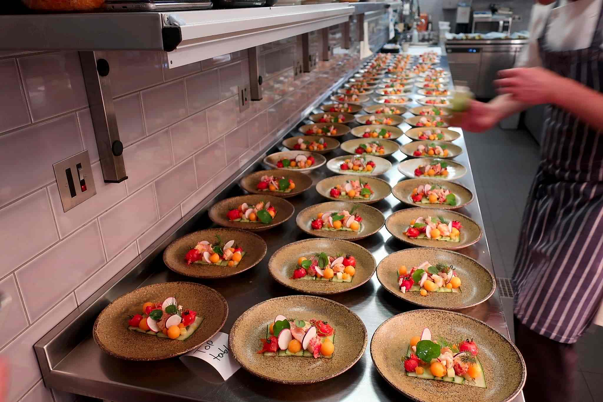 Hurley House - A chef arranges plates of colorful gourmet salads on a long kitchen counter. Each plate features a mix of vegetables, greens, and edible flowers, prepared for a large event or restaurant service.