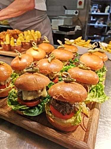 Hurley House - A wooden tray of mini burgers with lettuce, tomato, and melted cheese. Each burger is topped with a small green pepper and secured with a skewer. In the background, there are small bowls of golden fries.