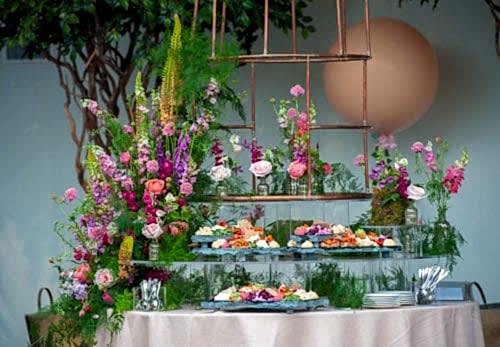 Hurley House - A beautifully arranged floral display with vibrant pink, purple, and green flowers surrounds a tiered tray of colorful, assorted appetizers on a table. In the background, theres a large peach-colored balloon and lush greenery. Plates and cutlery are on the side.