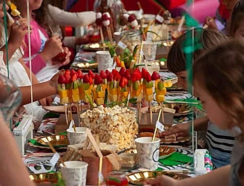 Hurley House - Children are gathered around a festive table, enjoying a colorful assortment of fruit skewers and popcorn. The table is decorated with vibrant patterns and set with drinks and gold plates, creating a lively and joyful atmosphere.
