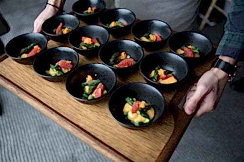 Hurley House - A person holds a wooden tray with multiple small black bowls containing a colorful dish. Each bowl appears to have greens, slices of red fruit, and an orange garnish, presented on a light gray carpeted floor.
