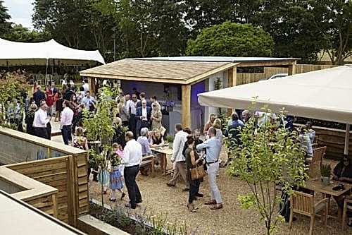 Hurley House - A lively outdoor gathering with people mingling around tables in a garden setting. Theres a wooden structure in the center, trees and tents in the background, and various individuals are engaged in conversation and socializing.