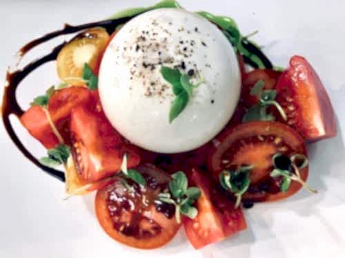 Hurley House - A plate of Caprese salad featuring a ball of fresh mozzarella garnished with cracked pepper and basil leaves, surrounded by slices of red and yellow tomatoes. Drizzles of balsamic glaze and olive oil add flavor and visual appeal.