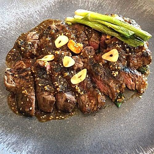 Hurley House - A plate of grilled steak slices topped with sesame seeds and garlic slices, accompanied by spring onions. The dish is garnished with a savory sauce, presented on a dark, textured plate.