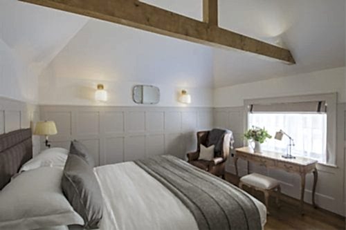 Hurley House - A cozy bedroom with a white bed, gray blanket, and wooden beams. Theres a leather armchair, desk, and stool near a window with a blind. Two wall sconces and a small mirror add lighting and decor to the paneled walls.