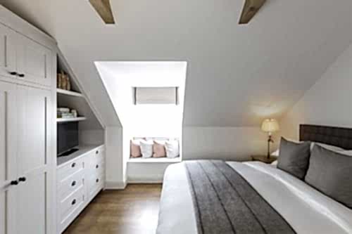 Hurley House - A cozy bedroom with white walls and wooden beams features a neatly made bed with gray accents, a window seat with cushions, a white wardrobe with shelves containing a TV and baskets, and a bedside lamp. Natural light fills the room.