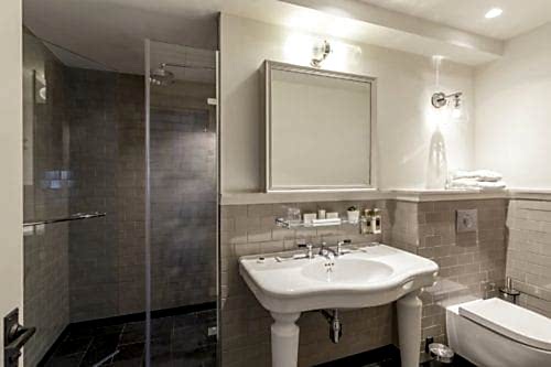 Hurley House - Modern bathroom with a white sink on a pedestal, a large mirror above, and a shower with a glass door on the left. Beige tiles cover the walls, and a white toilet is on the right. Shelves hold towels and toiletries, and the lighting is bright.